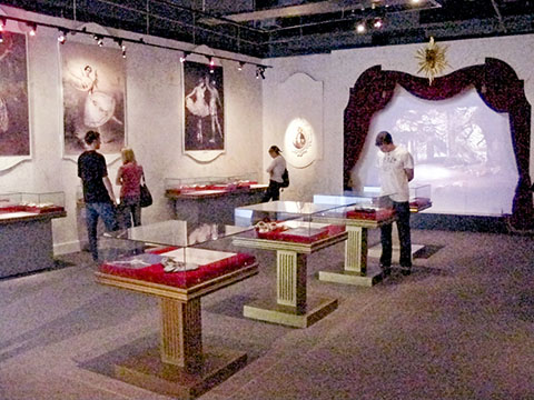 The exhibit space is dressed to create the feeling of classical ballet. Posters of famous dancers adorn the walls. A screen on a simulated stage projects video clips and display cases exhibit the history and craft of the ballet shoe.